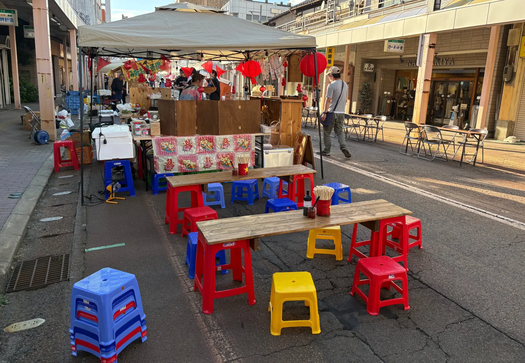 商店街のイベントで、屋台のテントの前に赤・青・黄色のプラスチックスツールと木のテーブルを並べた様子